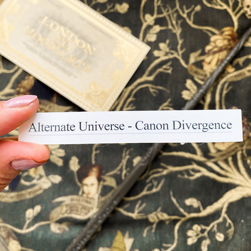 Photo of a white and black sticker in the style of Archive of Our Own fanfiction tags. Reads 'Alternate Universe - Canon Divergence'. Measures approximately 4 by 2 inches.