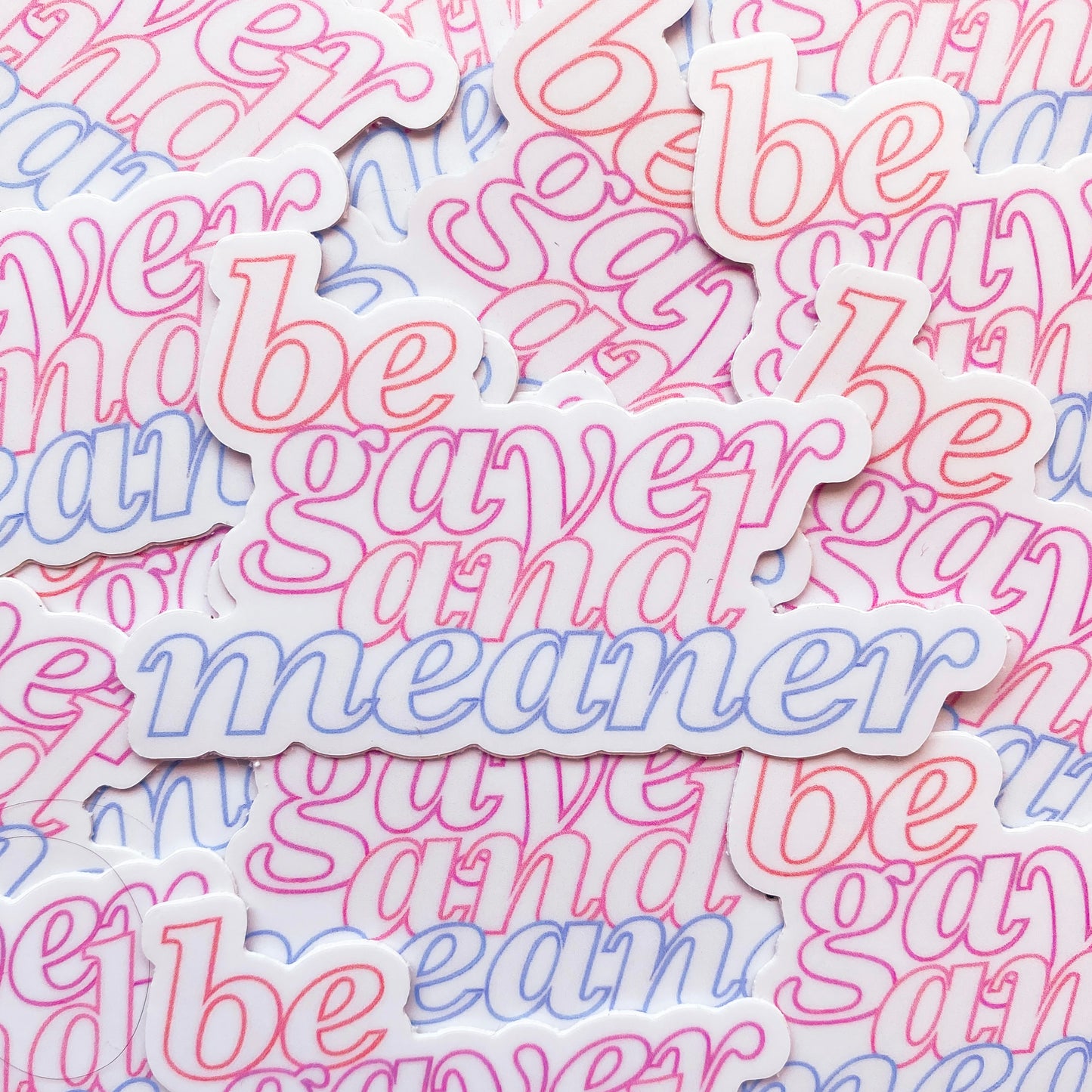 Photo of a white sticker with the words 'be gayer and meaner' in pride flag colours of red, orange, pink and blue.